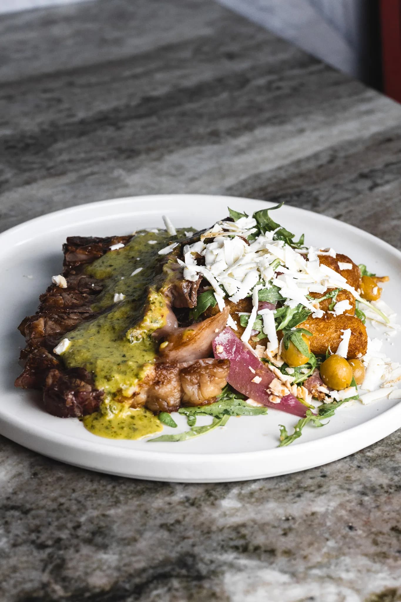 Sliced grilled steak topped with herb sauce, with crispy fried sides and shaved cheese on a stone counter.