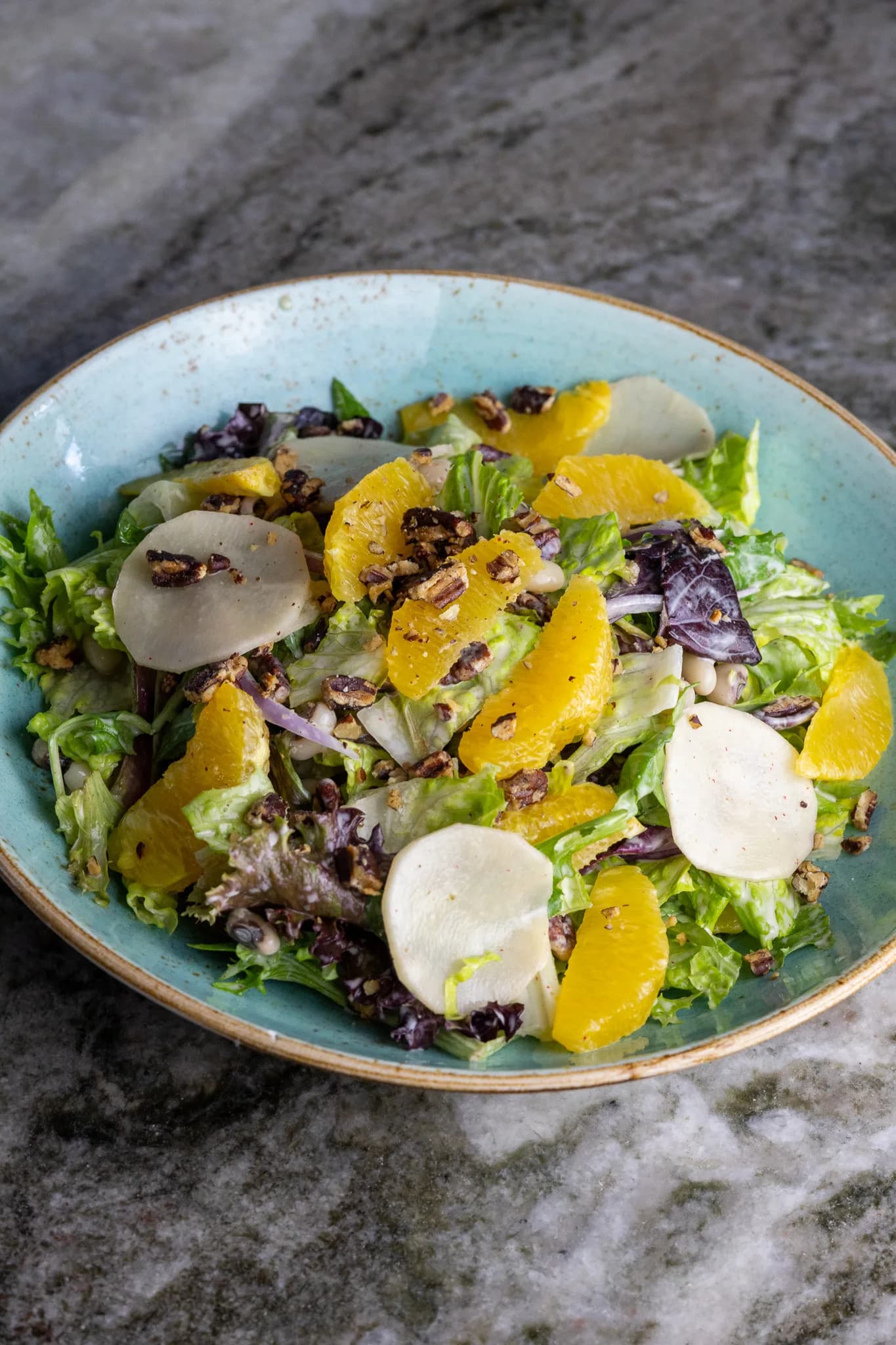 A teal bowl of mixed greens with citrus segments, candied pecans, and shaved root vegetables.