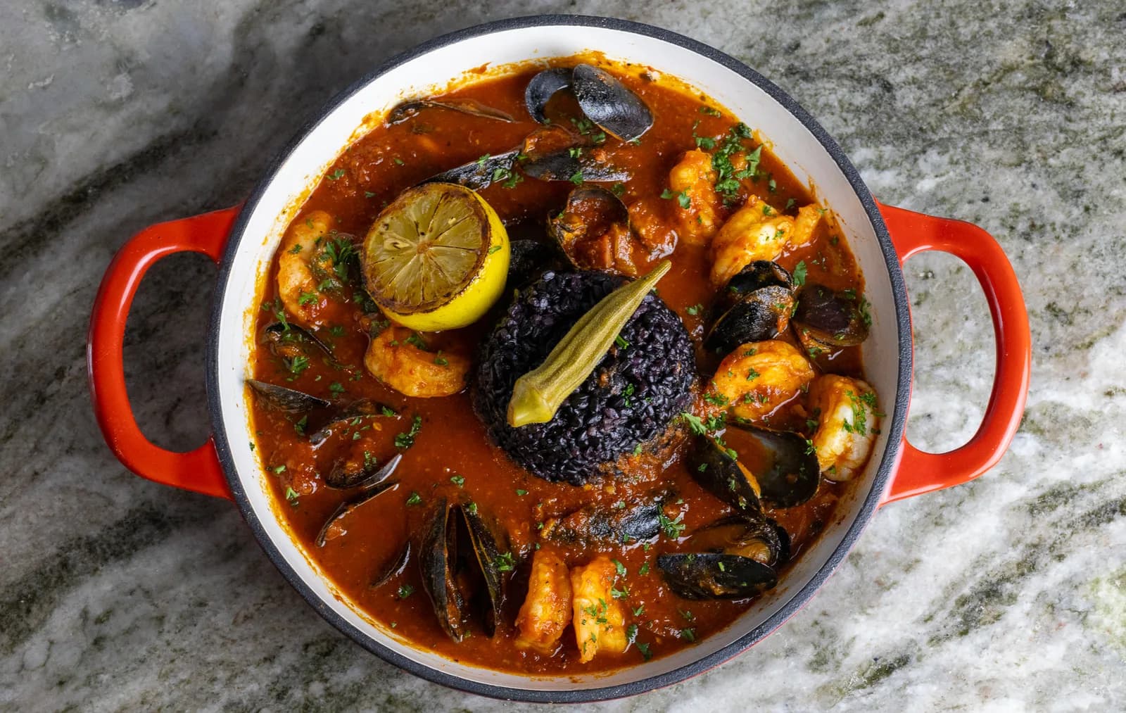 A red enameled pot of seafood stew with mussels, shrimp, charred lemon, herbs, and a mound of black rice.